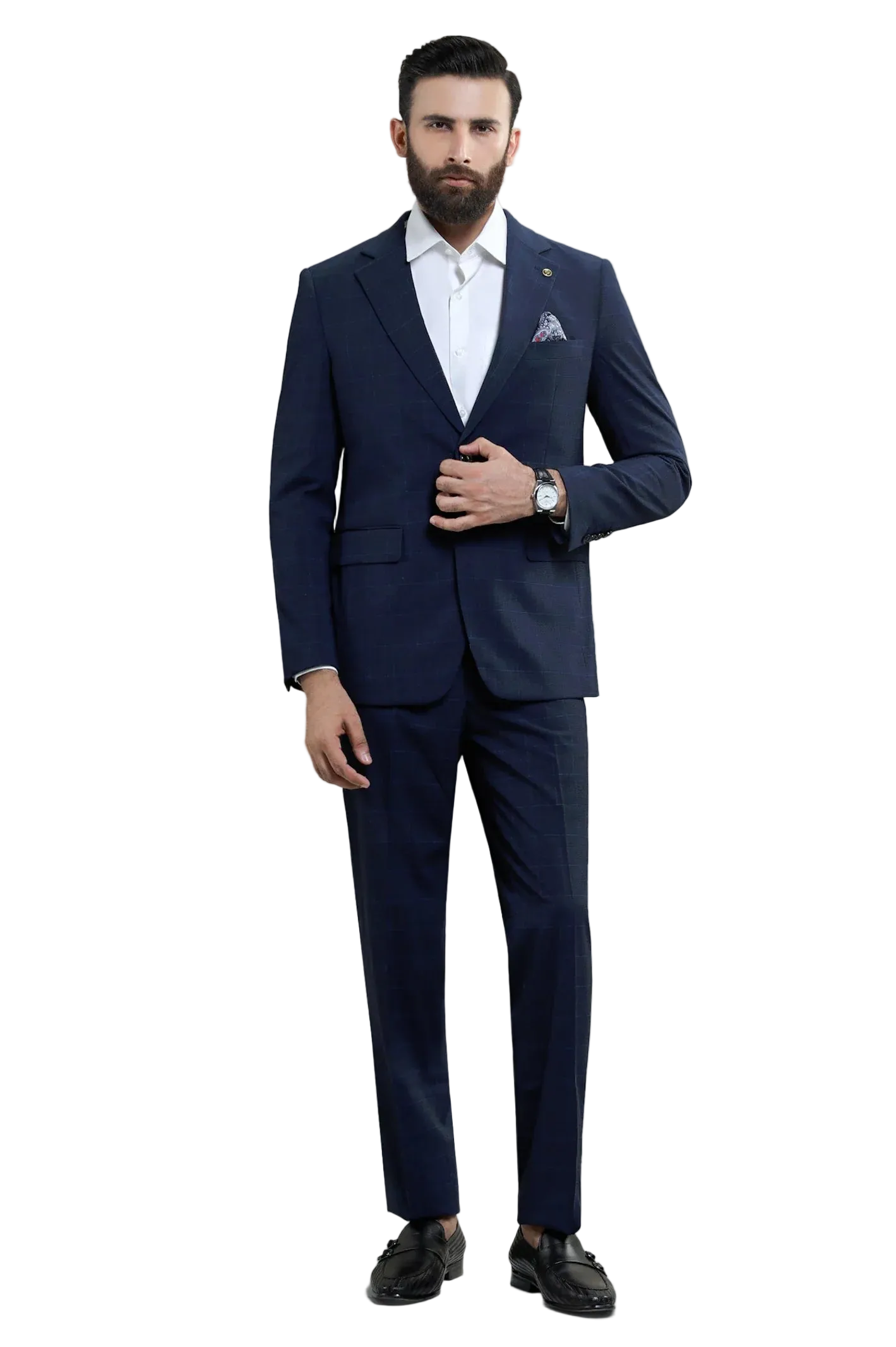 A man wearing a navy blue two-piece suit with a white dress shirt, a patterned pocket square, and dark loafers, standing against a light gray studio background.